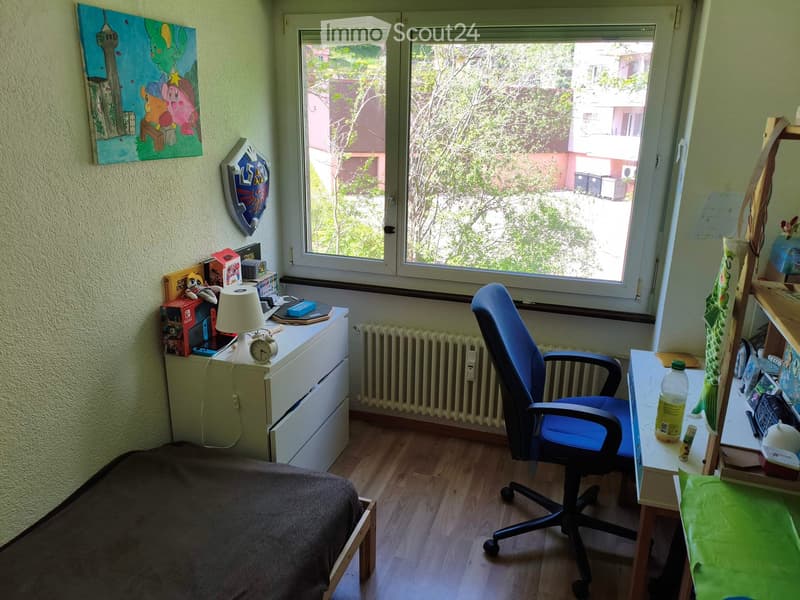 A small, cozy bedroom with a bed, a desk with a chair, a lamp, and various items including books and a gaming console. The room has wooden flooring and large windows offering a view of the outside.