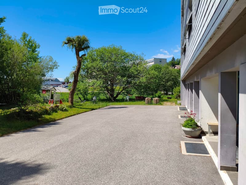 A paved driveway leads to a building with a small garden area featuring trees, chairs, and a planter with flowers.
