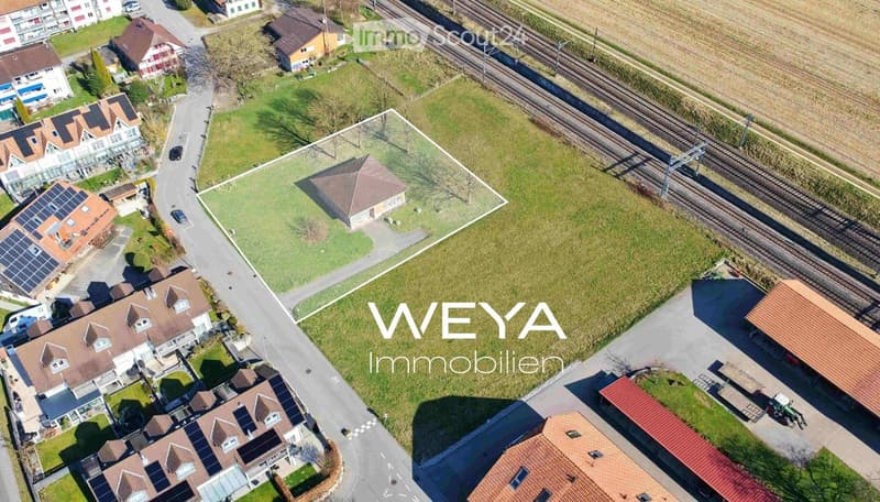 Aerial view of a residential area with multiple houses, solar panels, a railway line, and surrounding greenery.