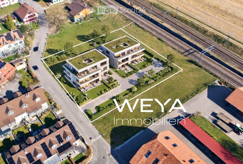 Aerial view of a modern residential complex with green roofs, surrounded by neighboring houses, a road, and a railway track in the background.