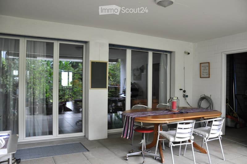 Balcony with a round wooden table, four chairs, and a candle on the table. Large sliding glass doors open to a lush garden area.