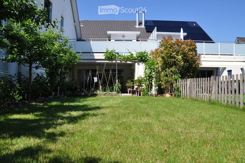 A well-maintained garden with a lawn, trees, and a wooden fence, adjacent to a modern house with a balcony and solar panels on the roof.