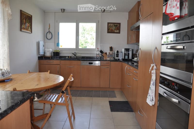 Bright kitchen with wooden cabinets, granite countertops, and stainless steel appliances including an oven and dishwasher. Features a round wooden dining table with chairs, a sink with a window above, and various kitchen items on the counter.