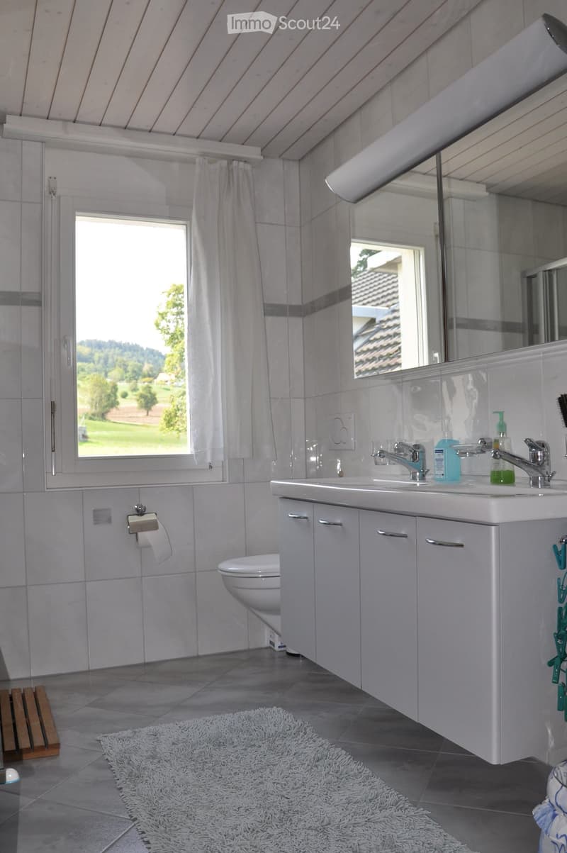 Modern bathroom with white tiles, a large window, a double sink with two faucets, a toilet, a mirror, and a fluffy white rug on the floor.