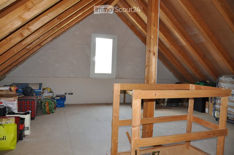 An attic space with wooden beams, a window, and a partially constructed wooden frame. Various items such as bags, boxes, and a fan are scattered around.