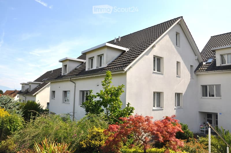 A modern two-story house with white exterior walls, dark grey tiled roof, multiple windows, and a small balcony area visible on the right side.