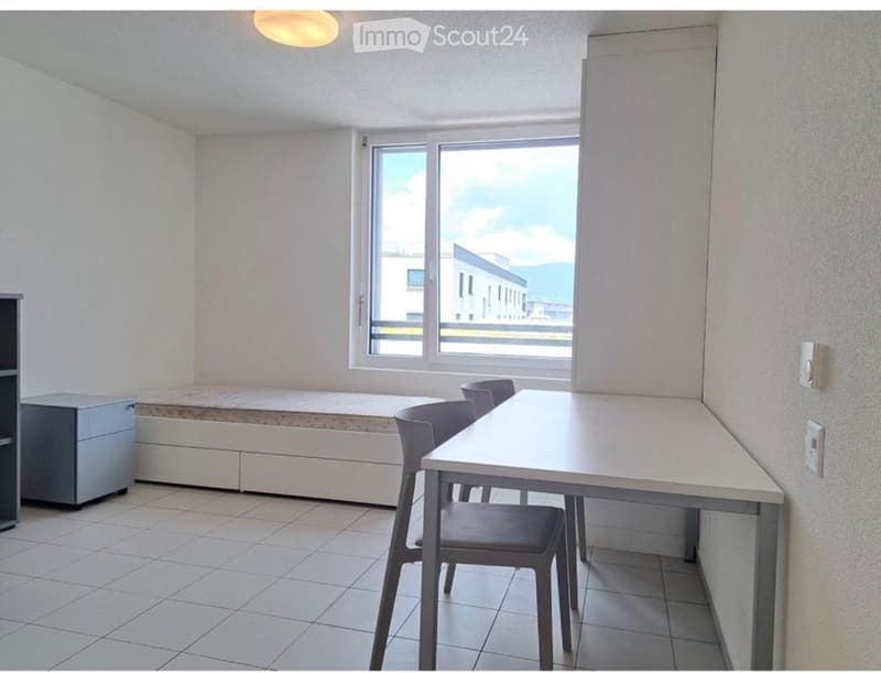 A small, furnished room with a bed, desk, and chair. The room has tiled flooring, white walls, and a large window offering a view of the outside.