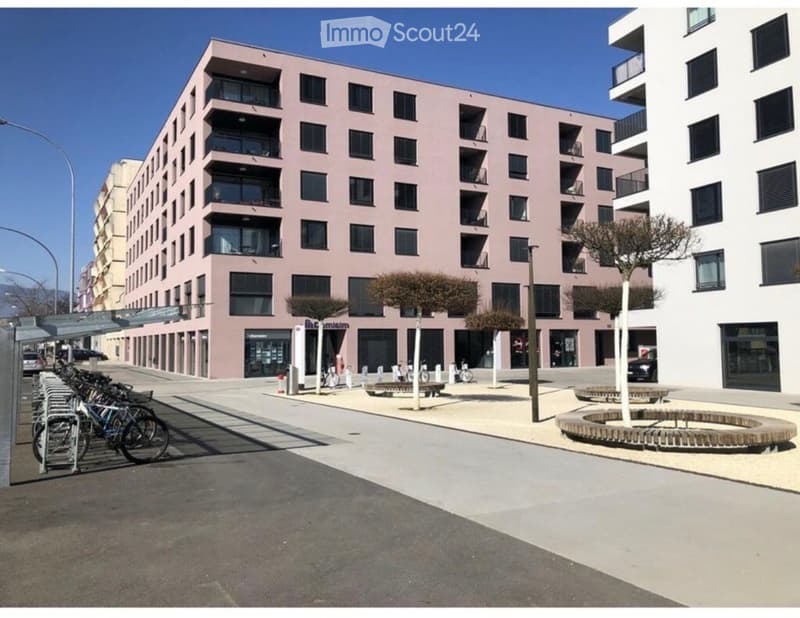 Modern multi-story building with pink facade and balconies, surrounded by a paved area with benches, trees, and bicycle racks.