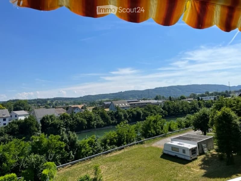Balcony with a scenic view of a river, surrounding greenery, and distant hills under a clear blue sky.