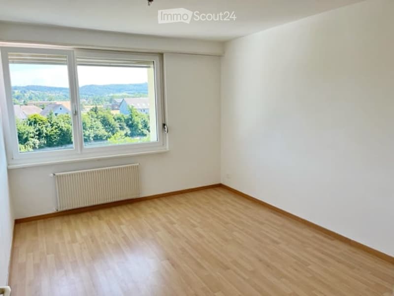 Empty room with wooden flooring, white walls, large window offering a scenic view, and a radiator below the window.