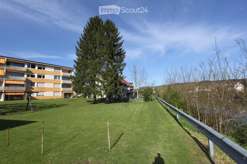 Well-maintained lawn area with a swing set, surrounded by trees and a multi-story building with balconies and orange accents.