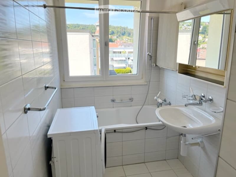 A small bathroom with a bathtub, sink, mirror, and radiator. The room has white tiled walls and floor, with a window offering a view of the surrounding area.