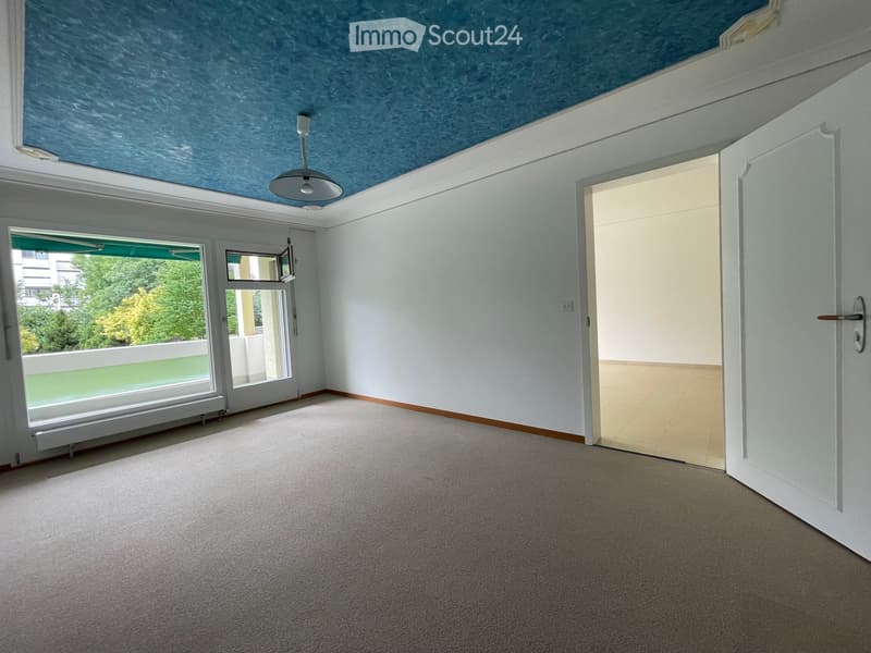 Empty room with white walls, carpeted floor, large window with view, and a door leading to another space.