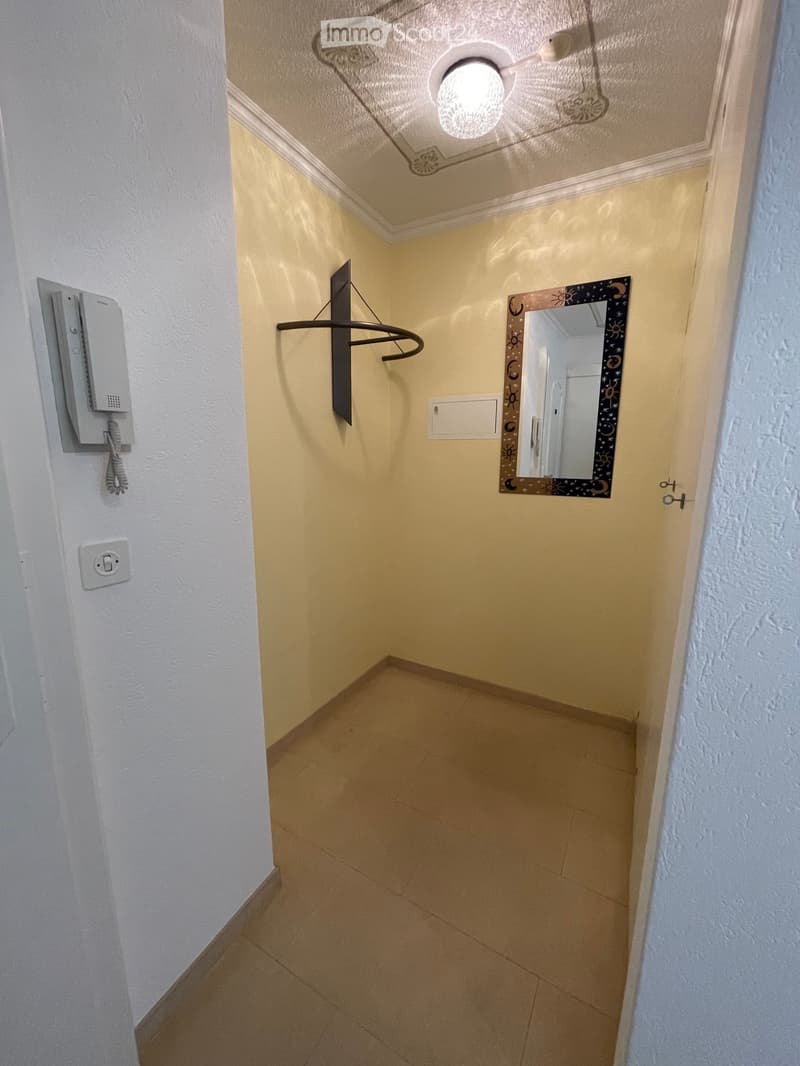 Narrow hallway with beige walls, tiled floor, and a decorative mirror with sun and moon designs. A coat rack and telephone are mounted on the walls.