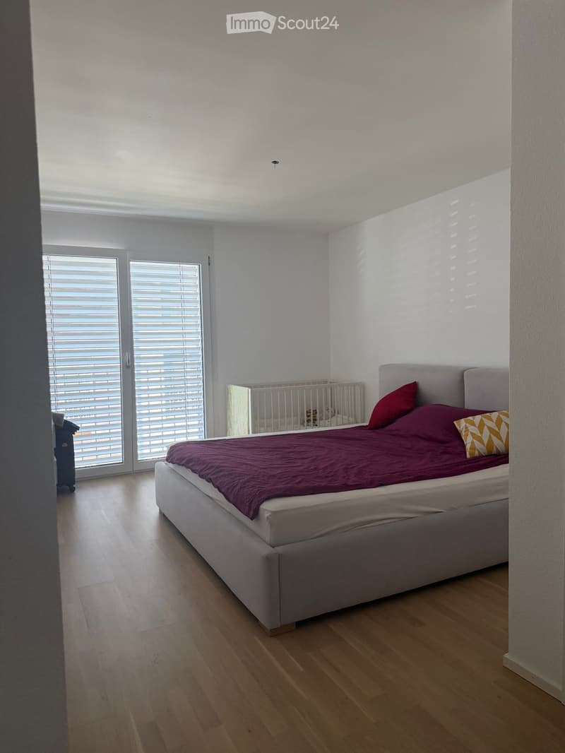 Bright bedroom with wooden flooring, large window with blinds, a neatly made bed with a purple duvet and decorative pillows, and a small crib with a toy inside.