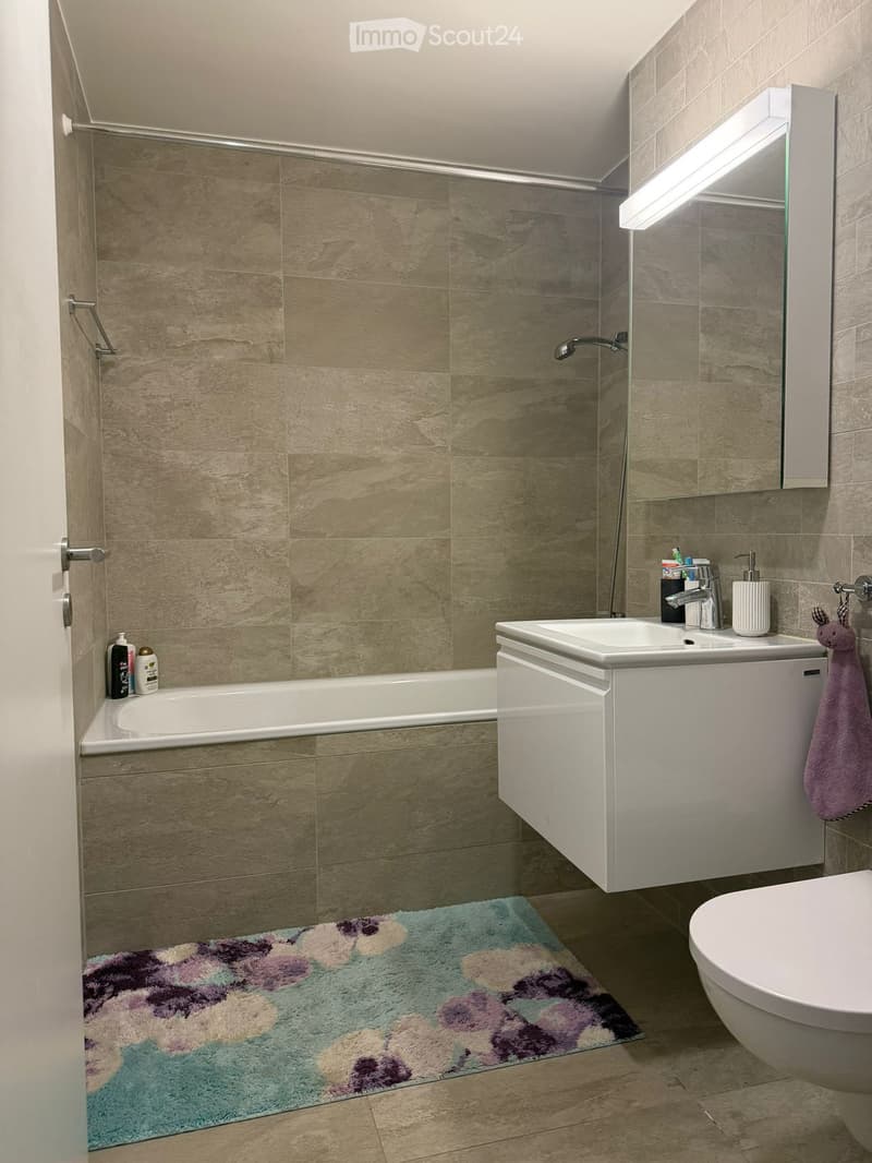 Modern bathroom with beige tiled walls, a bathtub, a sink with a modern faucet, a toilet, a mirror with built-in lighting, a colorful bathroom rug, and a towel hanging on a rack.