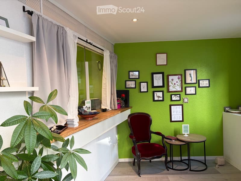 A small, well-lit room with green walls, a red upholstered chair, a small round table, a potted plant, and several framed pictures on the wall.