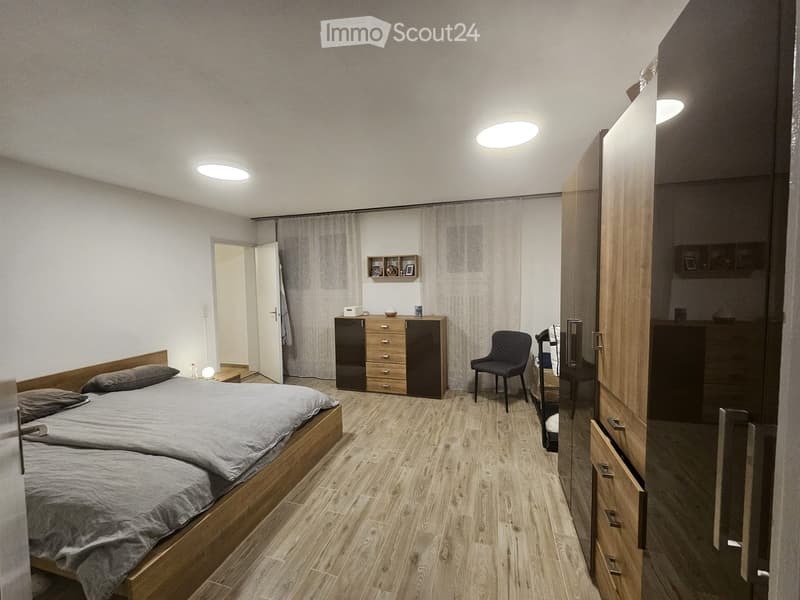 Modern bedroom with wooden flooring, a double bed with grey bedding, a wardrobe, a nightstand, a chair, and a shelf with decorative items.