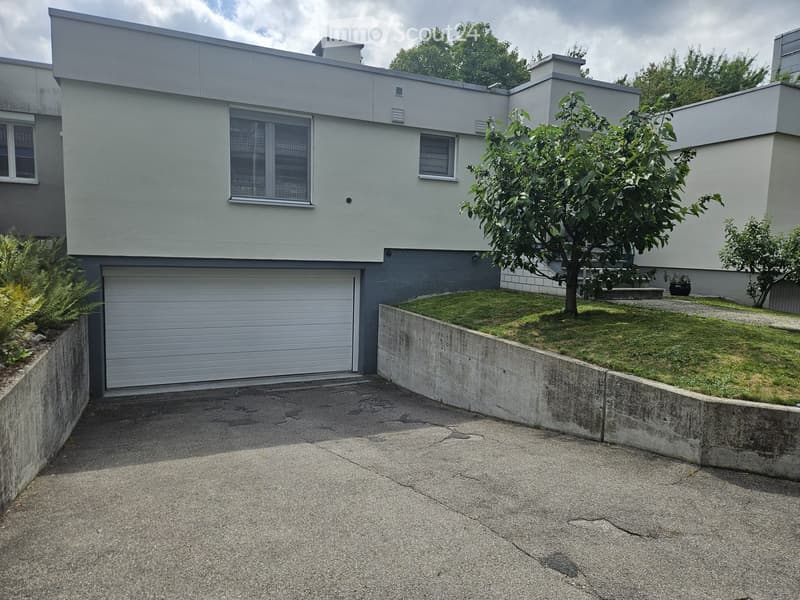 Modern two-story house with a flat roof, large windows, and a garage. The house is surrounded by a low concrete wall and a small garden with a tree and shrubs.