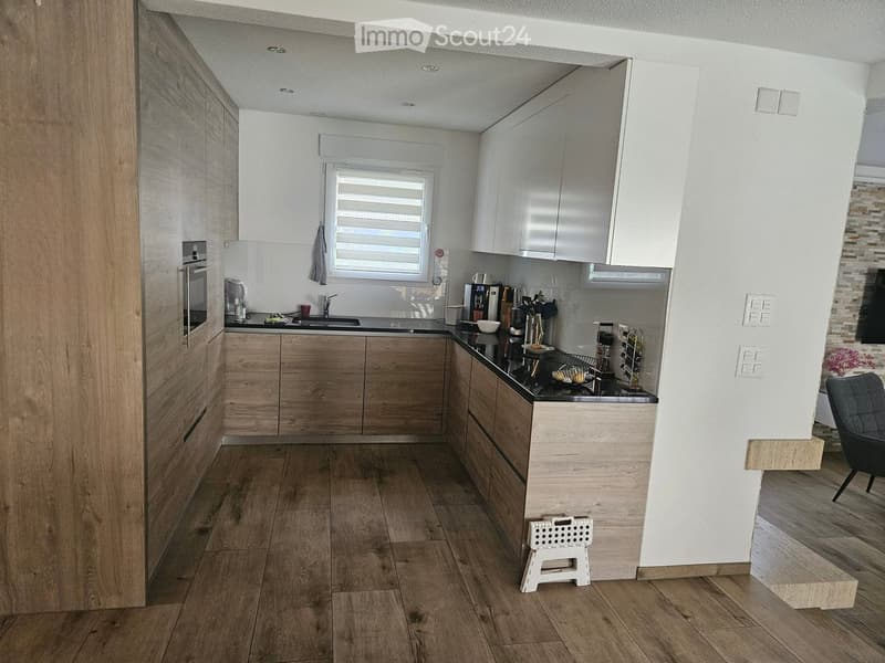 Modern kitchen with wooden cabinets, stainless steel appliances, and a black countertop. Features include a sink, coffee maker, and a small white stool near the island.