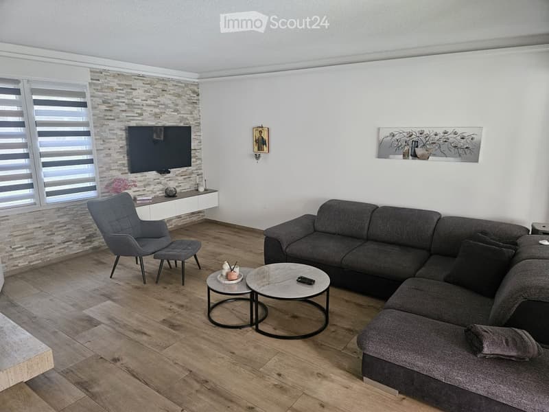 Modern living room with a grey sectional sofa, armchair, two round coffee tables, TV mounted on a stone accent wall, and decorative artwork.