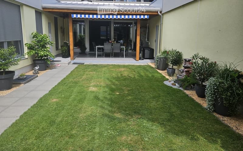 Well-maintained garden with a neatly trimmed lawn, surrounded by potted plants and a covered outdoor seating area with chairs and a table.