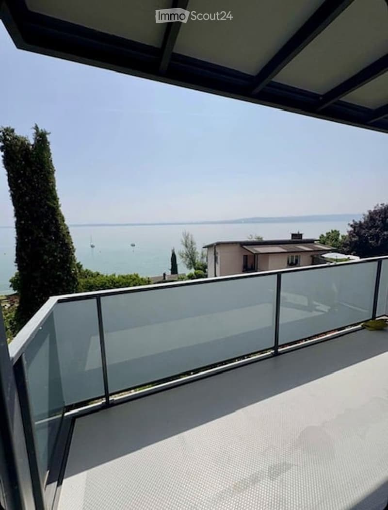 A balcony with a glass railing and a view of the sea and a house in the distance.