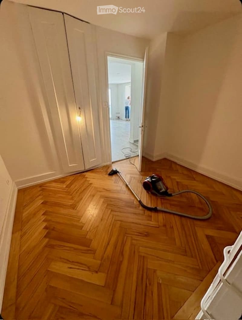 Empty room with wooden parquet flooring, white walls, and an open door leading to another room. A vacuum cleaner is on the floor.