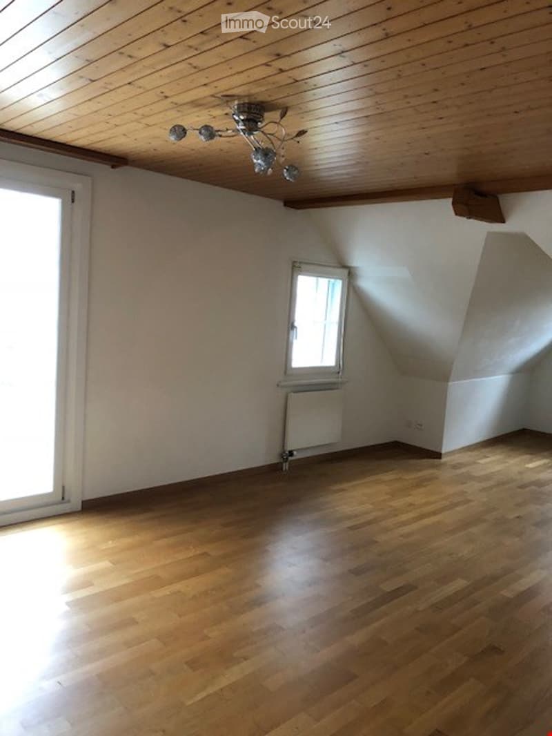 Empty room with wooden flooring, white walls, and a slanted wooden ceiling. Features include a large window, a smaller window, a radiator, and a chandelier light fixture.