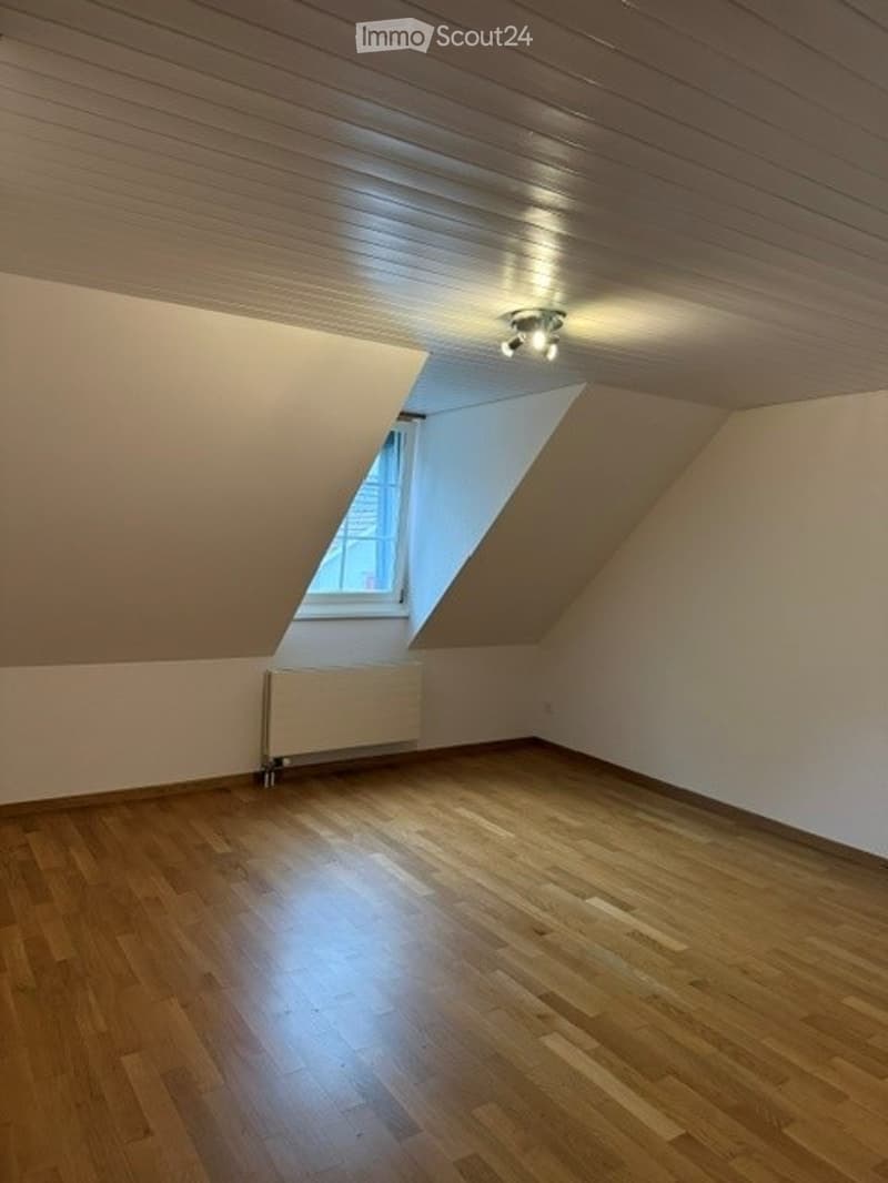 Empty attic room with wooden flooring, slanted wooden ceiling, single window, and a radiator. No furniture or equipment present.