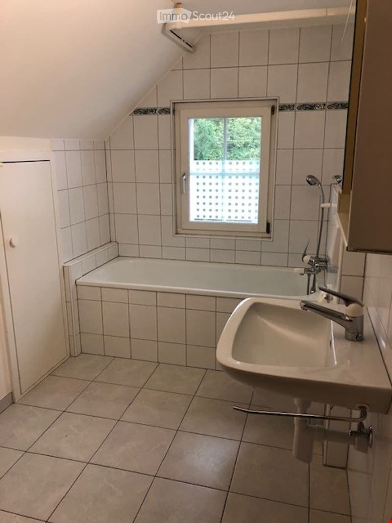 Bathroom with a bathtub, sink, and window with a view of greenery outside. The room features white tiled walls and a slanted ceiling typical of an attic space.