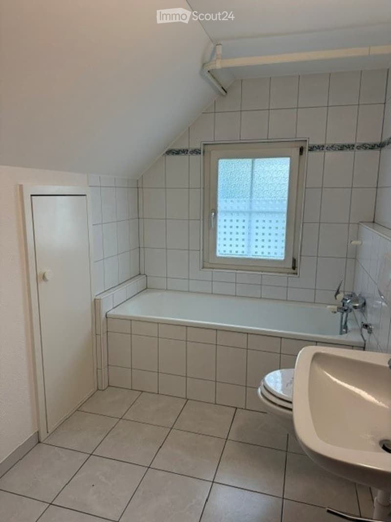 Bright bathroom with white tiled walls and floor, featuring a bathtub, sink, and toilet. A window with frosted glass provides natural light.