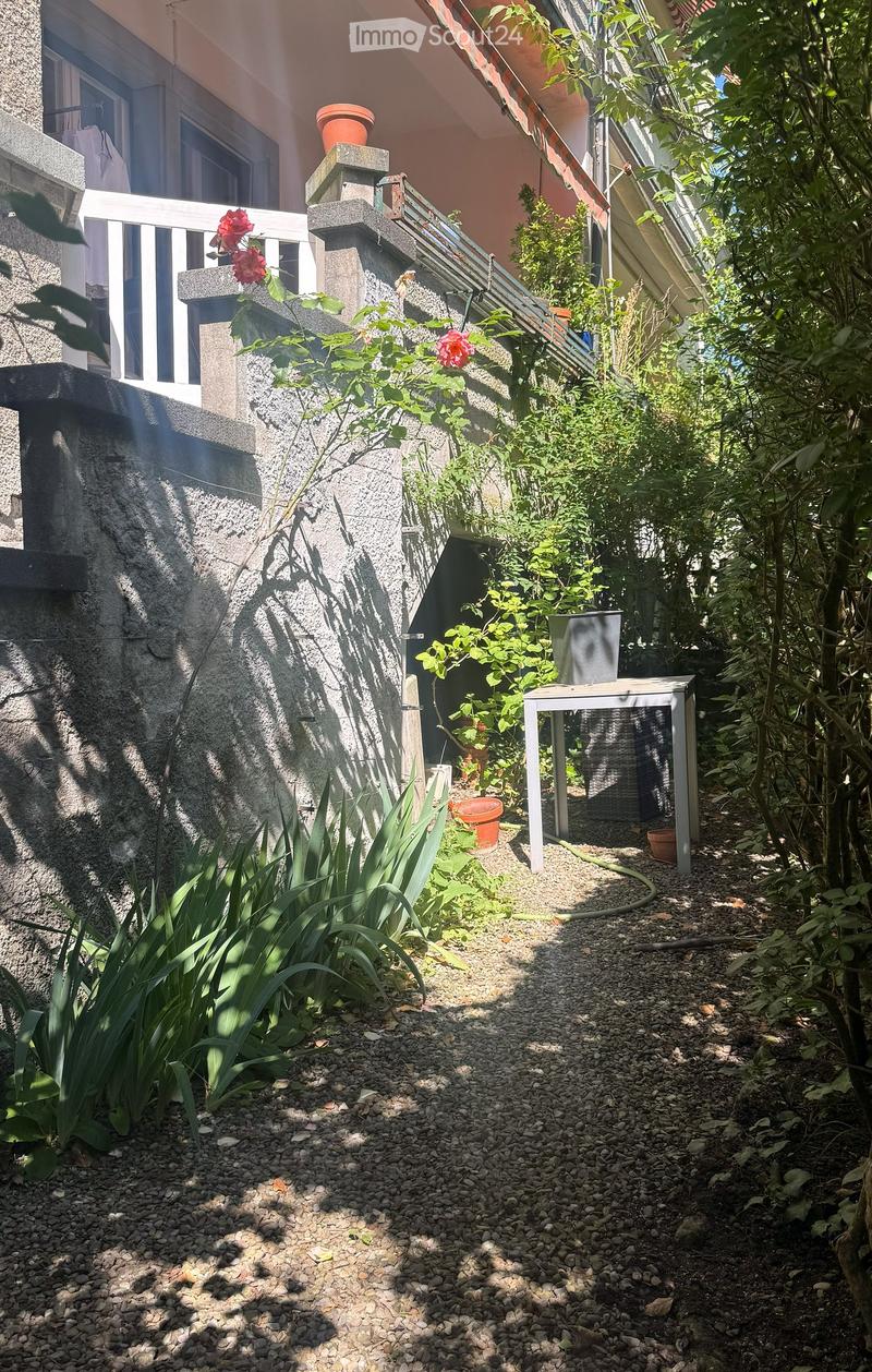 Garden area with gravel ground, various plants, small table, and potted flowers near a building staircase.