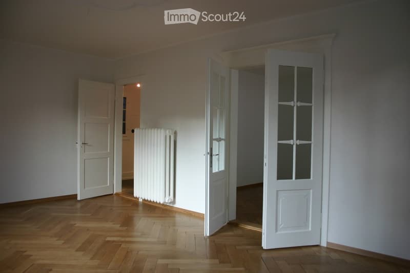 Empty room with wooden flooring, white walls, and two open doors leading to adjacent spaces. A white radiator is visible against one wall.