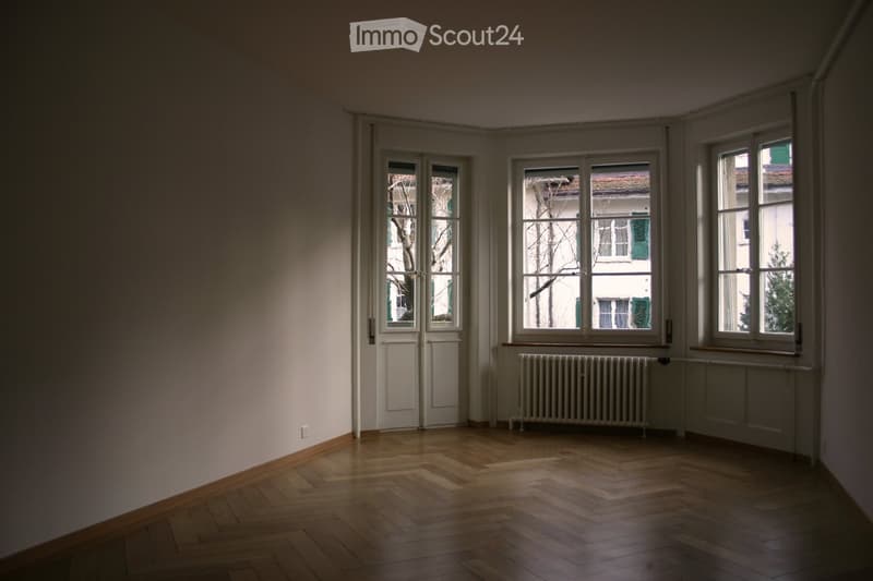 Empty room with wooden flooring, large windows, radiator, and a door leading outside.