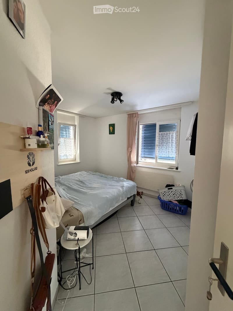 A small bedroom with a bed, a bedside table, a laptop, a handbag, and a laundry basket. The room has tiled flooring, white walls, and two windows with curtains.
