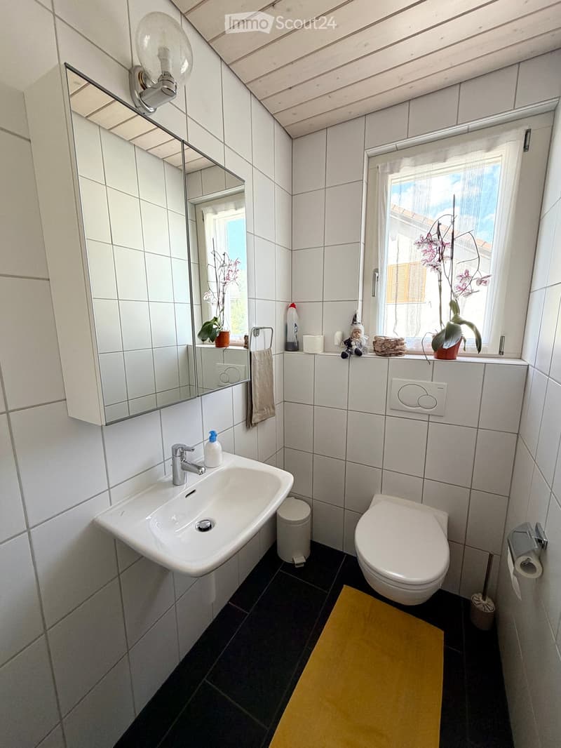 Modern bathroom with white tiles, a sink, toilet, mirror, towel, and small decorative items on the windowsill.