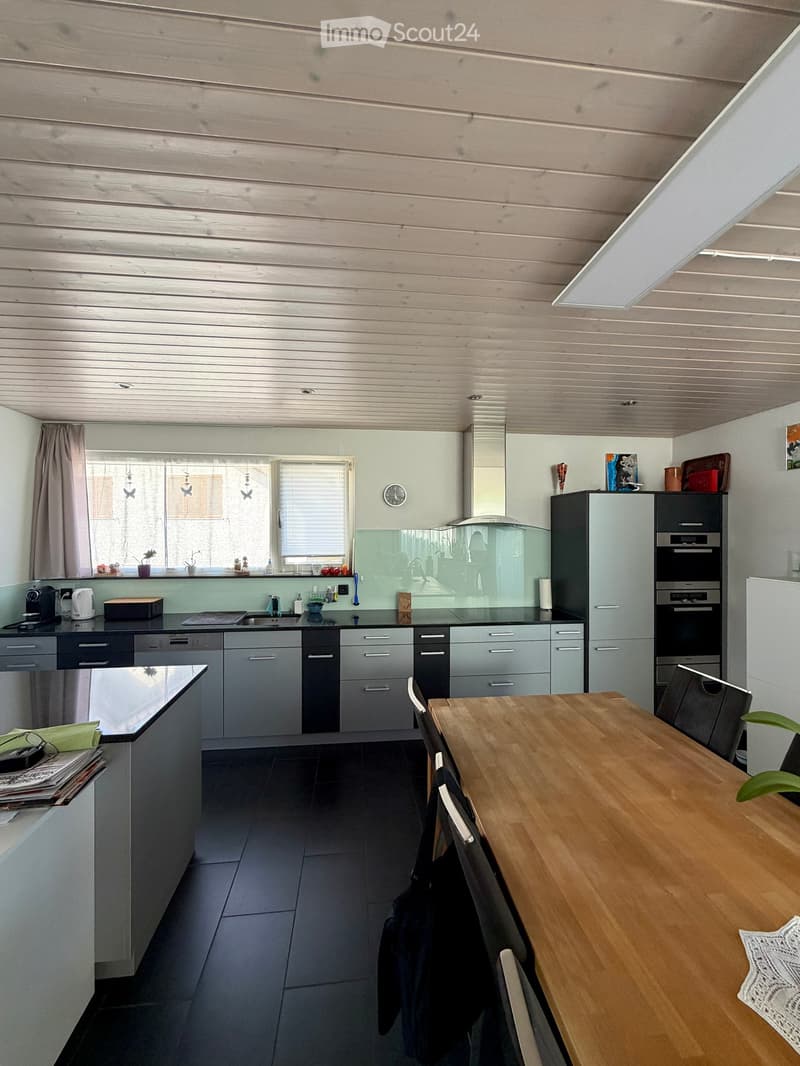Modern kitchen with grey cabinets, stainless steel appliances, wooden dining table, and black tiled floor.