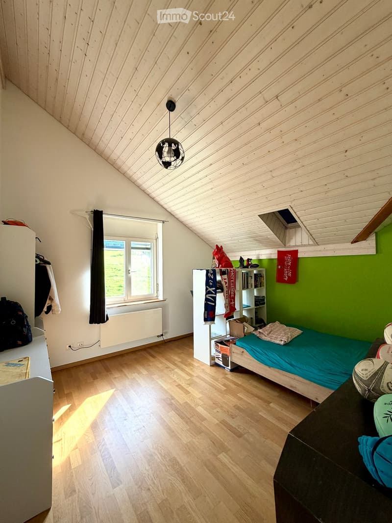 Cozy attic bedroom with wooden floors, slanted wooden ceiling, large window with curtain, bed with blue bedding, bookshelf, and various personal items.