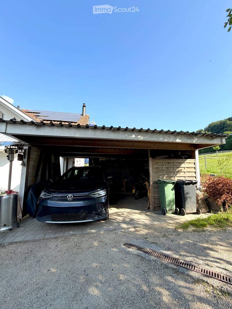 Garage with a car parked inside, covered by a wooden roof, surrounded by a paved area with trash bins and a small garden area.