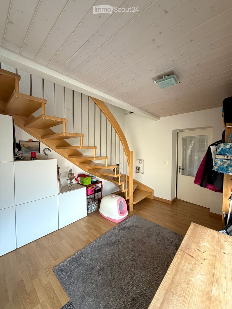 A modern hallway with a wooden staircase, a cat litter box, a small table with various items, and a grey rug on the floor.