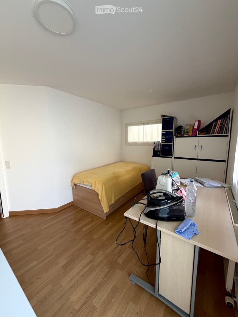 A small, furnished room with a bed, a chair, a table with an iron and other items, a cabinet, and a shelf with books and other items.