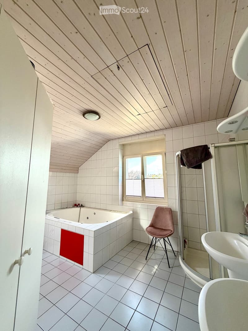 Bathroom with white tiled walls and floor, a bathtub with a red towel, a sink, a pink stool, and a shower area with a towel hanging on the side.