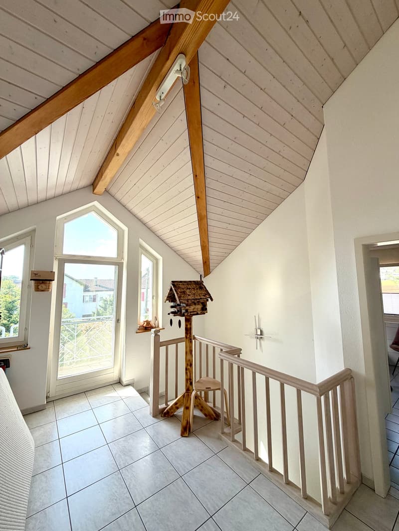Bright hallway with white walls, wooden ceiling beams, and a staircase leading upwards. Features include a bird feeder on a stand, a small stool, and windows offering a view of the outdoors.