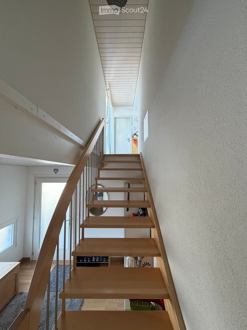 Modern wooden staircase with metal railing, leading to an upper level. The walls are painted in a light neutral tone, and there is a small window on the left side providing natural light. A shoe rack is visible at the bottom of the stairs, and a door is s