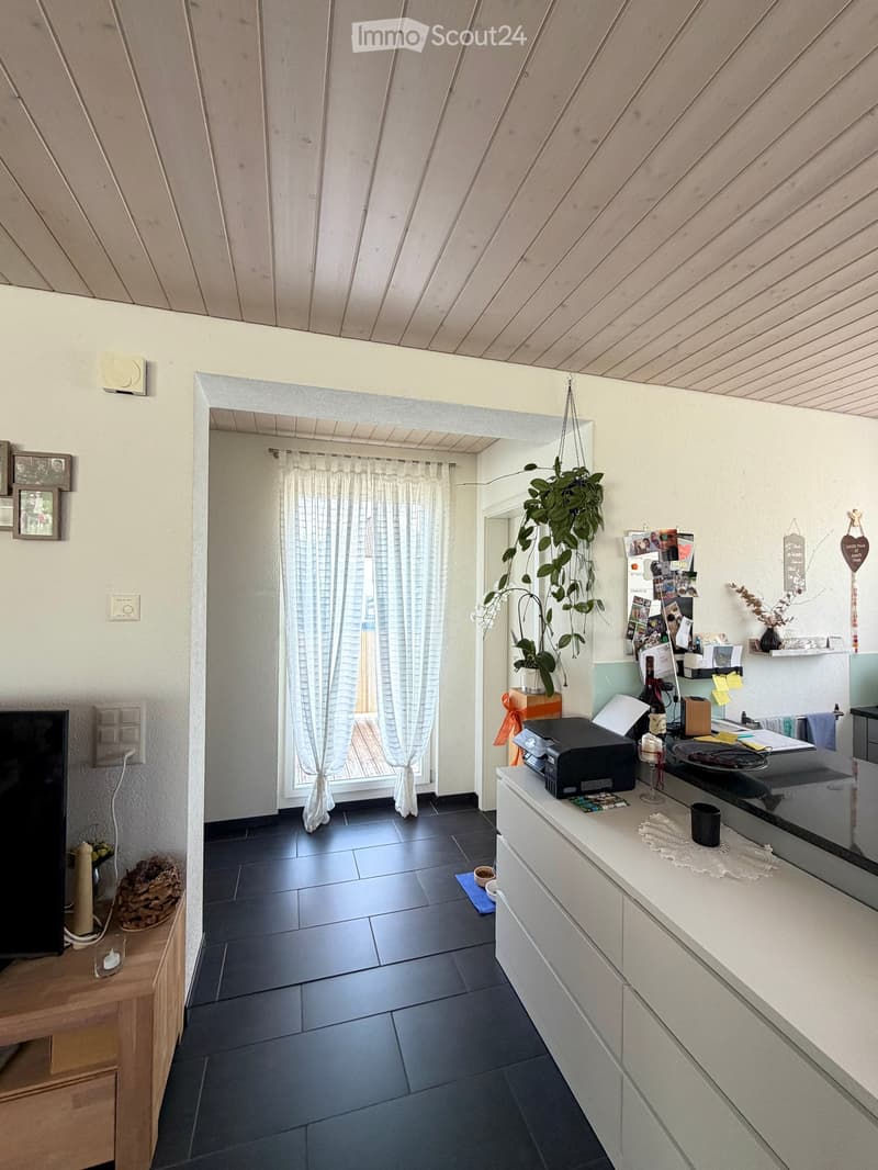 A modern, well-lit room with a television on a wooden stand, a printer on a white counter, and a hanging plant. The room features a large window with white curtains, a dark tiled floor, and various decorative items including picture frames and a woven bas