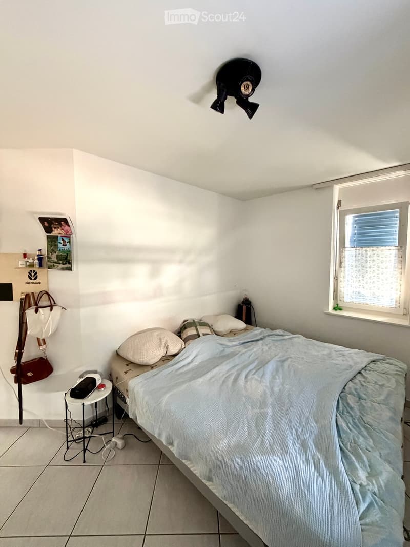 A small, neatly arranged bedroom with a single bed, pillows, a blue bedspread, a nightstand with a lamp, and a handbag hanging on the wall. The room features white walls, a tiled floor, and a window with blinds.