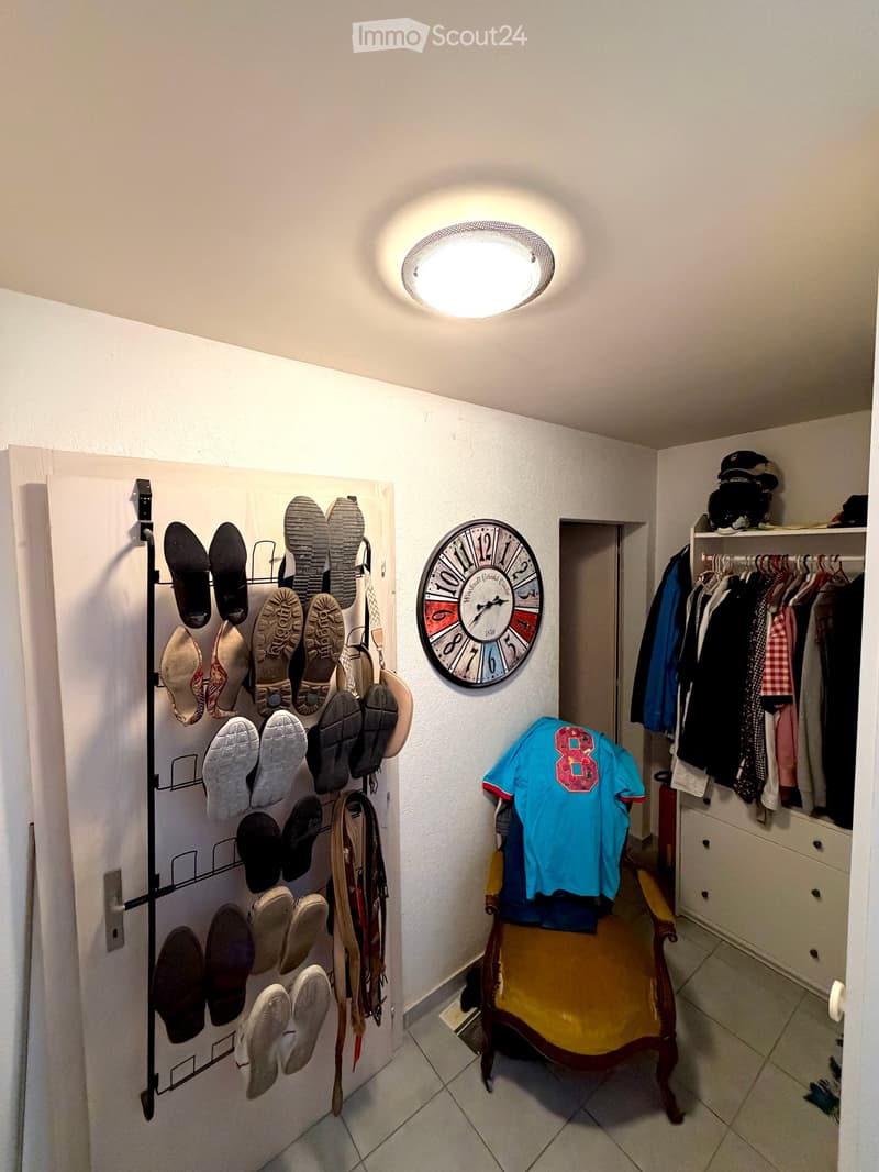 Hallway with shoe rack, clock, clothing rack, and a small chair with a shirt draped over it.