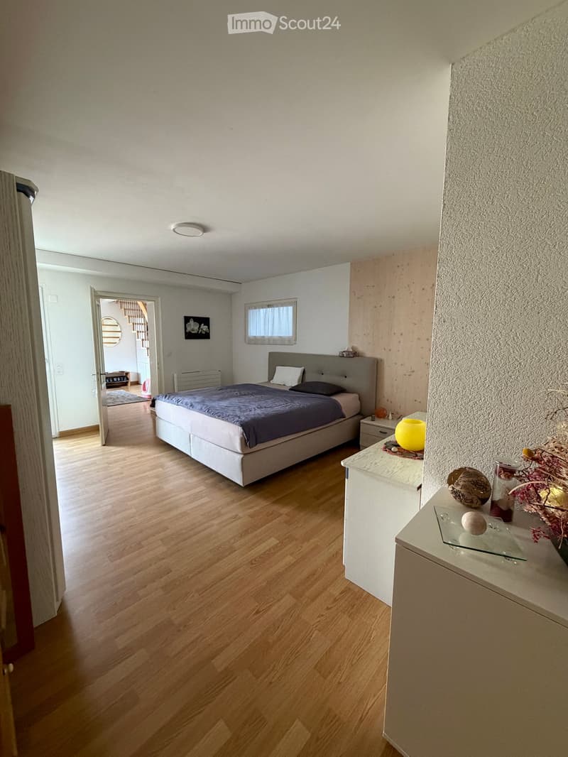 Modern bedroom with wooden flooring, a large bed with dark bedding, a nightstand with a lamp, and a small table with decorative items.