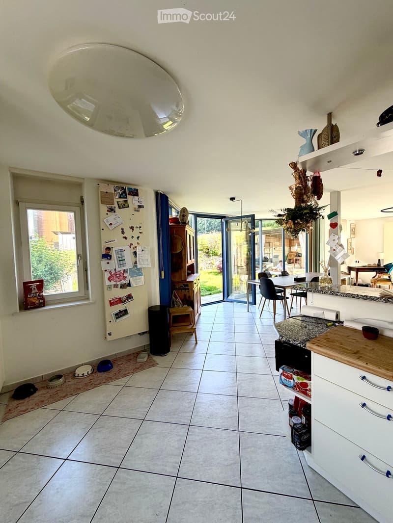 Bright kitchen with tiled floor, white cabinets, granite countertop, and a dining area with a table and chairs. A large window and sliding glass doors lead to a garden. A refrigerator with notes and magnets is visible, along with a small stool and dog bow